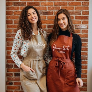 Pu&ograve; includere: Due donne con grembiuli. Uno &egrave; beige con la scritta "Wedding APRON" in bianco. L'altro &egrave; color ruggine con la scritta "Your text CUSTOM" in bianco. Entrambi i grembiuli hanno tasche e sono legati in vita.