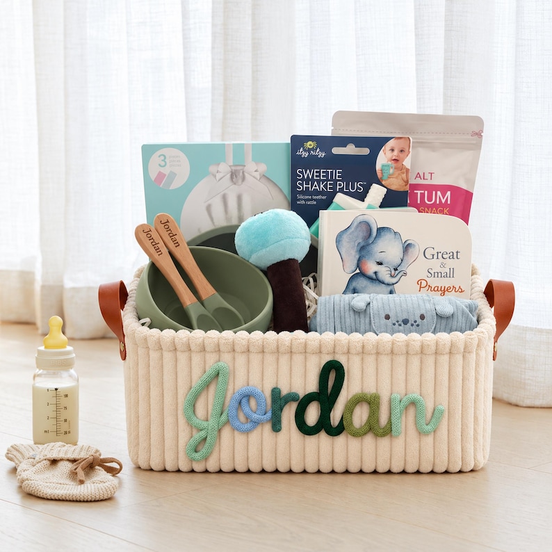 May include: A cream-colored corduroy storage basket with the name Jordan in multi-colored yarn lettering. Inside are a green silicone bowl, two wooden spoons, a blue plush toy, a book titled Great & Small Prayers, and baby items.