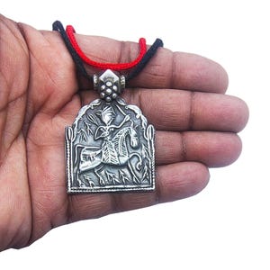 May include: A silver pendant necklace featuring a detailed relief of a person on horseback, with a red and black cord. The pendant has an ornate, rectangular shape with a decorative border. The necklace is held in a hand.
