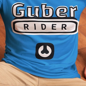 Può includere: Una maglietta blu con la scritta "Guber RIDER" in bianco e nero. Sotto il testo c'è una grafica in bianco e nero. La maglietta è a maniche corte e la persona indossa pantaloni color cachi.