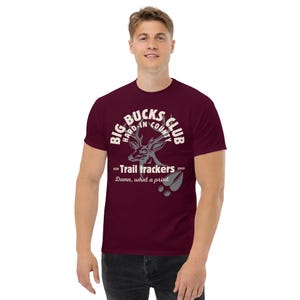 May include: A maroon t-shirt with a graphic design featuring a deer head and the text "BIG BUCKS CLUB HARD-IN COUNTY" in a circular arrangement. Below, it reads "Trail trackers 2025" and "Damn, what a print."