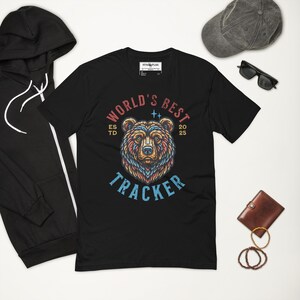 May include: Black t-shirt featuring a bear graphic with the text "WORLD'S BEST TRACKER". The design includes the text "ESTD 2025". A black hoodie, a grey cap, sunglasses, a brown wallet, and rings are also visible.