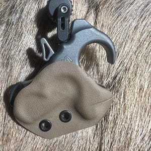 May include: A black and tan archery release aid with a curved handle and a tan leather-like grip. The release aid has a circular adjustment dial and two black eyelets on the grip.