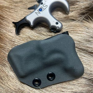 May include: A silver-colored archery release aid with the word "STAN" and a blue accent. A black trigger and a black, textured case with two eyelets are also visible. The items are displayed on a brown and tan fur-like surface.