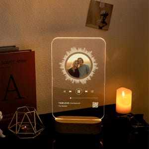 May include: A personalized acrylic night light with a photo of a couple in a circular design. The light displays a music waveform and song information, including the song title "Timeless" by The Weeknd. The base is made of wood.