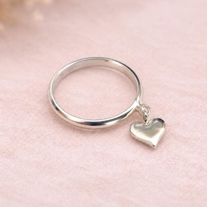 Puede incluir: Un anillo de tono plateado pulido que presenta un dije liso y sólido en forma de corazón que cuelga de una pequeña argolla circular unida a la banda principal. La superficie metálica refleja la luz, creando un aspecto brillante y metálico.