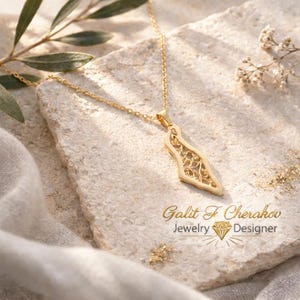 Könnte beinhalten: Eine goldfarbene Halskette mit einem Anhänger in Form der Landkarte Israels und einem filigranen Ausschnittmuster. Der Anhänger hängt an einer zarten goldfarbenen Kette. Der Text Galit F Cherakov Jewelry Designer ist sichtbar.