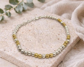 May include: Silver and gold beaded bracelet. The bracelet features alternating silver and gold-colored beads. The gold beads have a textured, faceted appearance. The bracelet is displayed on a light-colored stone surface with a soft, neutral-toned fabric in the background.