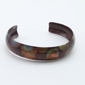 May include: A brown metal bangle bracelet with a colorful abstract pattern. The pattern features shades of green, orange, and blue.
