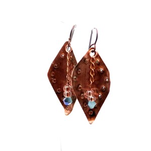 May include: A pair of copper earrings with a diamond shape and a textured surface. Each earring has a small chain with a clear bead and a blue crystal hanging from it.