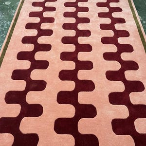 Può includere: Un tappeto rettangolare con un motivo ripetuto di linee ondulate rosso scuro su sfondo rosa chiaro. Il tappeto ha un bordo verde, che aggiunge un accento di contrasto. Adatto per soggiorno o camera da letto.