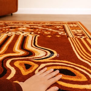 May include: A close-up of a brown rug with an abstract design in shades of orange, yellow, and black. The rug has a textured surface and a border with geometric patterns. The text "Rug Store" is visible on the rug.