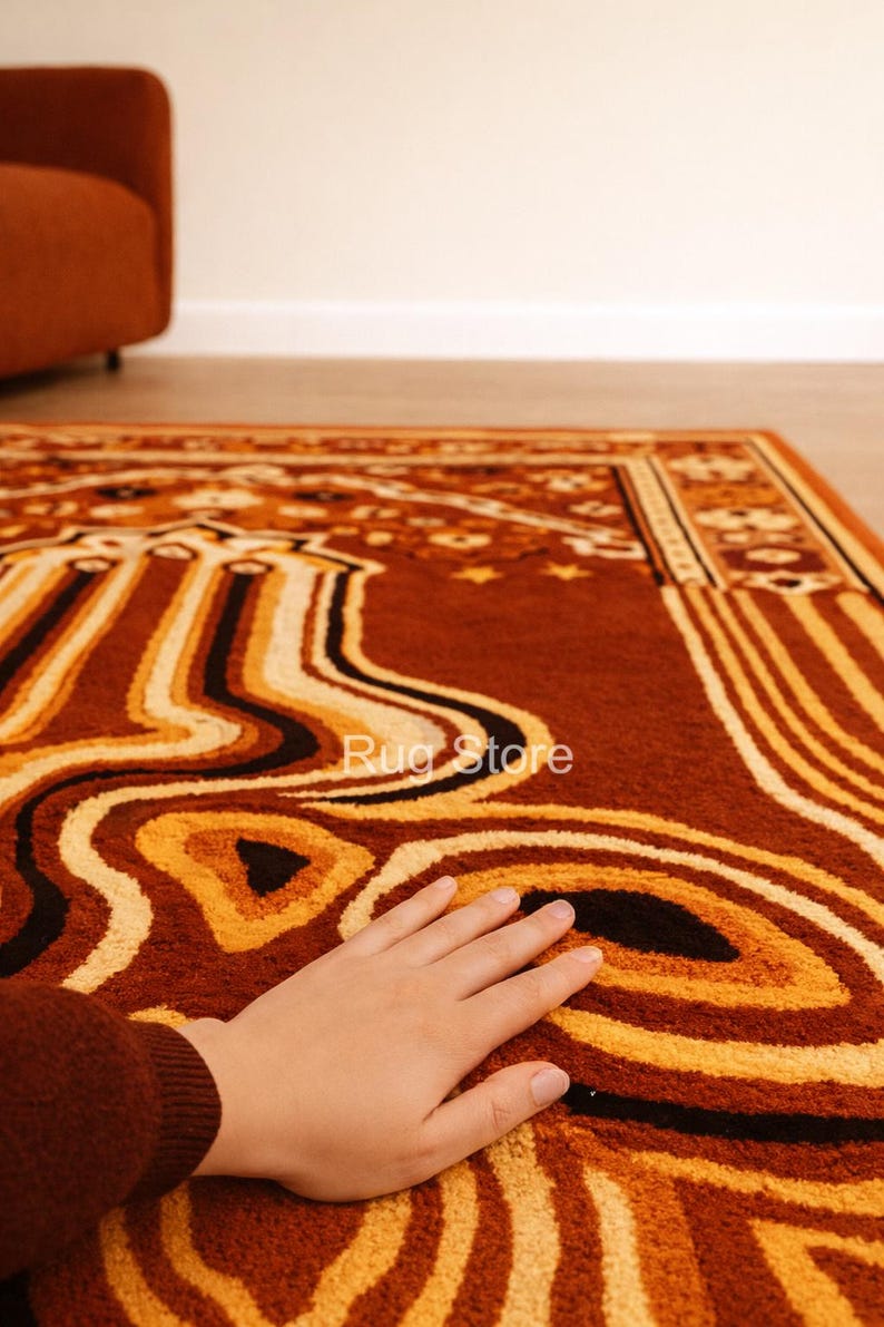 May include: A close-up of a brown rug with an abstract design in shades of orange, yellow, and black. The rug has a textured surface and a border with geometric patterns. The text "Rug Store" is visible on the rug.