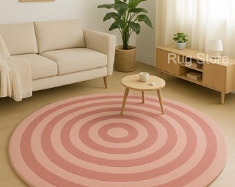 May include: A round rug with concentric circles in varying shades of pink. The rug is the focal point of a living room setting, with a sofa, side table, and cabinet visible. The words "Rug Store" are printed on the rug.