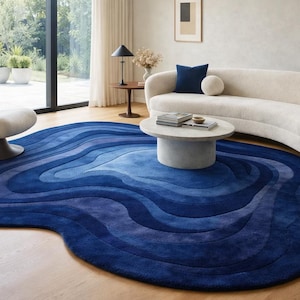 Könnte beinhalten: Ein modernes Wohnzimmer mit einem großen, blauen Teppich mit Wellenmuster. Das Design des Teppichs integriert Blautöne und erzeugt einen Farbverlauf. Ein geschwungenes weißes Sofa, ein runder Couchtisch und ein Drehstuhl vervollständigen die Einrichtung des Raumes.