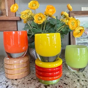 May include: Three colorful glass tumblers in orange, yellow, and green. The orange tumbler sits on a wooden base. The yellow tumbler is stacked on a base of red, orange, and green rings. Yellow flowers are in the background.
