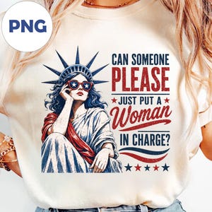 Könnte beinhalten: Ein cremefarbenes T-Shirt mit einer Grafik der Freiheitsstatue, die eine Sonnenbrille mit Sternenmuster trägt. Der Text lautet: CAN SOMEONE PLEASE JUST PUT A WOMAN IN CHARGE? Das Design verwendet rote, weiße und blaue Farben mit Sternakzenten.