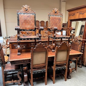 Peut inclure: Un ensemble de salle à manger comprenant une grande table en bois et plusieurs chaises. Deux chaises posées sur la table ont des dossiers hauts avec des panneaux en cuir brun gaufré et des cadres en bois foncé. Les autres chaises ont des dossiers en tissu.