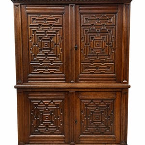 May include: A tall, dark brown wooden cabinet featuring four doors with intricate, geometric, pyramid-style carved patterns. The piece has a prominent overhanging top cornice, vertical frame detailing, and rests on four rounded, ball-shaped feet.