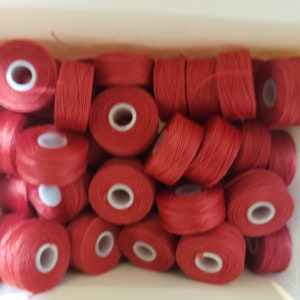 May include: A close-up of a box filled with spools of red thread. The spools are arranged in a random pattern.