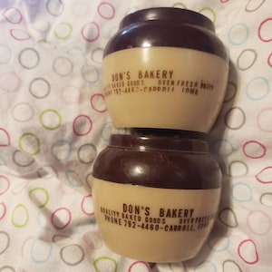 May include: Two brown and beige ceramic jars with a brown lid. The jars have the text "Don's Bakery" printed on them. The text also includes the phone number "792-4460" and the city "Carroll, Iowa".