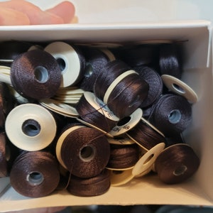 May include: A box filled with spools of brown embroidery floss. The spools are arranged in a random pattern and are all the same size. The spools are made of white plastic with a brown thread wound around them.