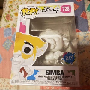 May include: A white, unpainted, Pop! Vinyl figure of Simba from Disney's The Lion King. The figure is in a clear plastic box with a colorful 'DIY' sticker. The box also features a colorful image of Simba on the front.
