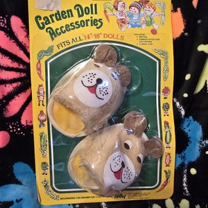 May include: A package of Garden Doll Accessories, featuring a pair of brown and yellow plush dog slippers for 14-18 inch dolls. The package includes a list of compatible dolls, including Softina, Cabbage Patch Kids, and Preemie Flower Kids. The package is yellow with green trim and features a picture of three dolls.