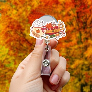 May include: A badge reel featuring a fall-themed design. The design includes a mug of whipped cream-topped beverage, a stack of books, a cup of tea, and autumn leaves. The reel has a clear plastic body and a metal clip.