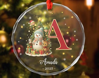 May include: A clear, round Christmas ornament with a red ribbon. The ornament features a snowman, penguin, and reindeer near a Christmas tree. A large red letter "A" is on the right, with the name "Amada" and the year "2025" below.