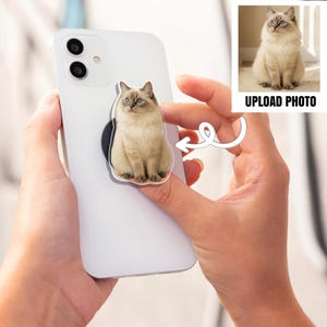 May include: A white smartphone with a pop-up grip featuring a Ragdoll cat with blue eyes. The cat is light brown with darker brown markings. The phone is held in a person's hands. The image also includes the text "UPLOAD PHOTO".