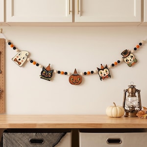 May include: A Halloween garland featuring felt balls in black, orange, and white, interspersed with wooden cutouts of a ghost, a black cat, a jack-o'-lantern, a devil, and a skull, all suspended on a string against a plain wall.