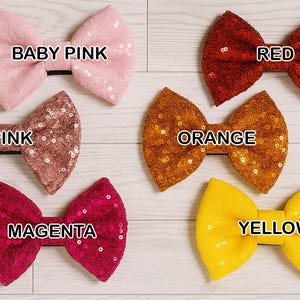 May include: A collection of six sequined bow ties in various colors: baby pink, pink, magenta, red, orange, and yellow. Each bow tie has a black band and is displayed on a light wood-grain surface. The color of each bow tie is labeled.