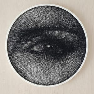 Op de afbeelding: Een cirkelvormig wandkunstwerk met een gedetailleerd oogontwerp, gemaakt met zwarte draad geweven over een wit frame. Het complexe draadkunstpatroon gebruikt variërende dichtheden van zwarte lijnen om de vorm van een iris, pupil en ooglid te vormen.