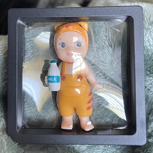 May include: A small, orange-yellow figurine of a baby wearing a tiger-themed hat and overalls, holding a milk bottle. The figurine is in a clear display case with a black frame. The word "MILK" is visible on the bottle.