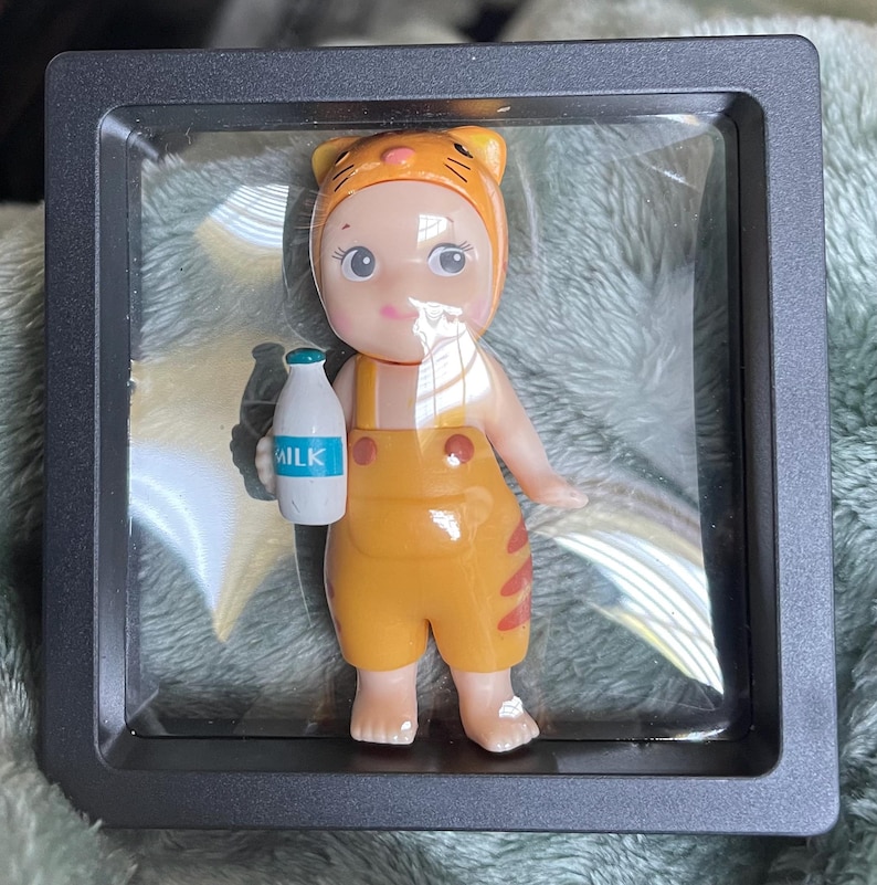 May include: A small, orange-yellow figurine of a baby wearing a tiger-themed hat and overalls, holding a milk bottle. The figurine is in a clear display case with a black frame. The word "MILK" is visible on the bottle.
