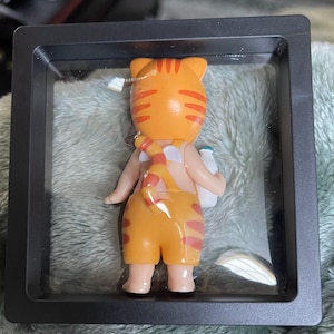May include: A cartoon cat figurine in an orange and red striped outfit, holding a white bottle. The cat is displayed in a black, square shadow box. The cat's tail is striped and wrapped around its back.
