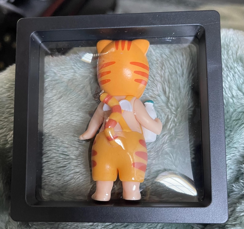 May include: A cartoon cat figurine in an orange and red striped outfit, holding a white bottle. The cat is displayed in a black, square shadow box. The cat's tail is striped and wrapped around its back.