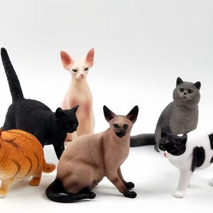 Può includere: Una collezione di sei statuette di gatti che rappresentano diverse razze e colori, tra cui un gatto tigrato arancione rotondo, un gatto nero slanciato, un gatto senza pelo rosa pallido, un siamese marrone e beige, un gatto grigio e uno maculato bianco e nero.
