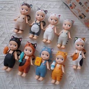 May include: A group of small figurines, each styled as a cat. The figures wear cat-themed outfits in colours such as black, orange, blue, and white, with matching cat ear hats and dungarees. Some hold miniature accessories like fish and milk bottles.