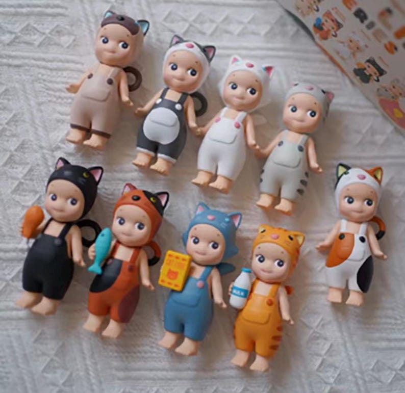 May include: A group of small figurines, each styled as a cat. The figures wear cat-themed outfits in colours such as black, orange, blue, and white, with matching cat ear hats and dungarees. Some hold miniature accessories like fish and milk bottles.