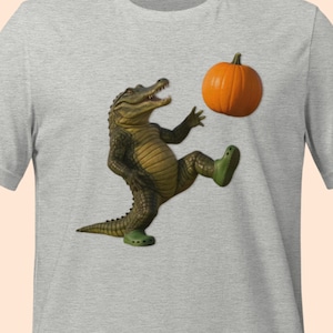 May include: A light gray t-shirt featuring a cartoon alligator kicking an orange pumpkin. The alligator is wearing green shoes. The design is centered on the shirt.