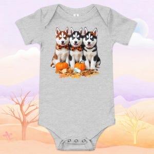 May include: A gray baby onesie featuring a graphic of three Husky puppies with bow ties, pumpkins, and autumn leaves. The puppies are brown, black, and white. The onesie has short sleeves and snap closures.
