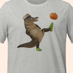 May include: A light gray t-shirt featuring a cartoon alligator wearing a fedora and green clogs, kicking a small orange pumpkin. The alligator is brown and tan with a detailed texture.