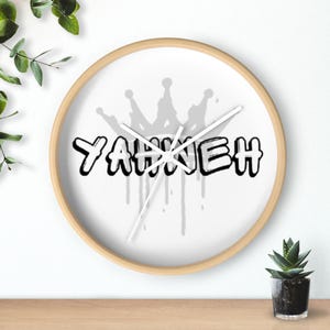 May include: A round wall clock with a light wood frame. The clock face is white with the word "YAHKEH" in black graffiti-style lettering, with a gray crown above and gray drips below. The clock hands are white.