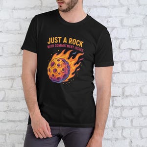 Może przedstawiać: Czarny t-shirt z kolorową, kreskówkową ilustracją meteoru w płomieniach. Nad grafiką znajduje się napis "JUST A ROCK WITH COMMITMENT ISSUES". Koszulka wykonana jest z miękkiego materiału.
