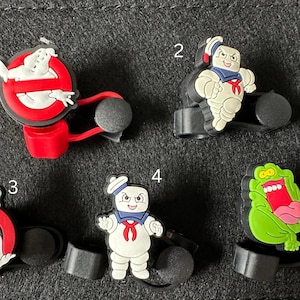 May include: A collection of five colorful shoe charms featuring characters from the Ghostbusters franchise. The charms include the Ghostbusters logo, Stay Puft Marshmallow Man, and Slimer, in various poses and designs.
