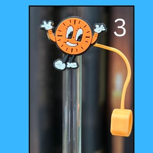 May include: Orange and black cartoon clock charm with white accents, arms raised, and a smiling face. Attached to a clear straw with an orange cap. The number 3 is visible in the upper right corner.