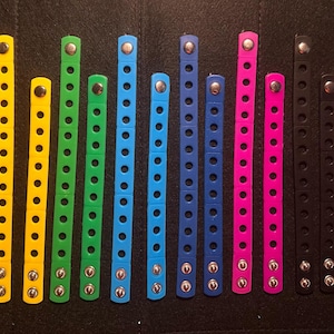 May include: A collection of colorful, adjustable bracelets in various shades, including red, orange, yellow, green, blue, pink, black, and white. Each bracelet has multiple holes and metal snap closures. The bracelets are arranged on a dark surface.
