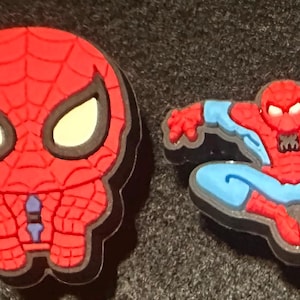 May include: Two red and blue Spider-Man shoe charms. One charm is a close-up of Spider-Man's head, and the other shows Spider-Man in a crouching pose. Both charms have a black border and are on a dark gray surface.